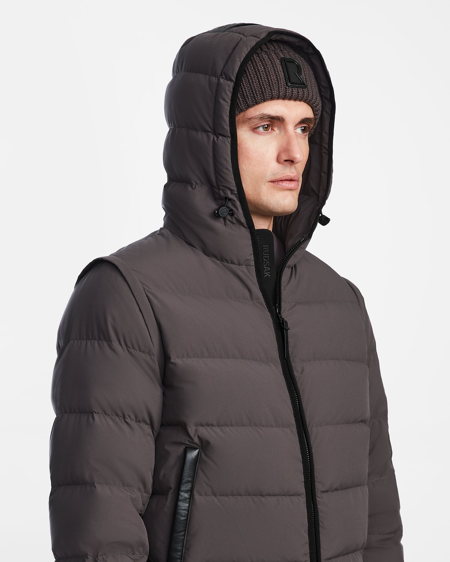 A man in the Rudsak DEMIAN M-R - M123504 Grey mens down puffer jacket, hood up and knit beanie on, looks to the side against a plain white background.