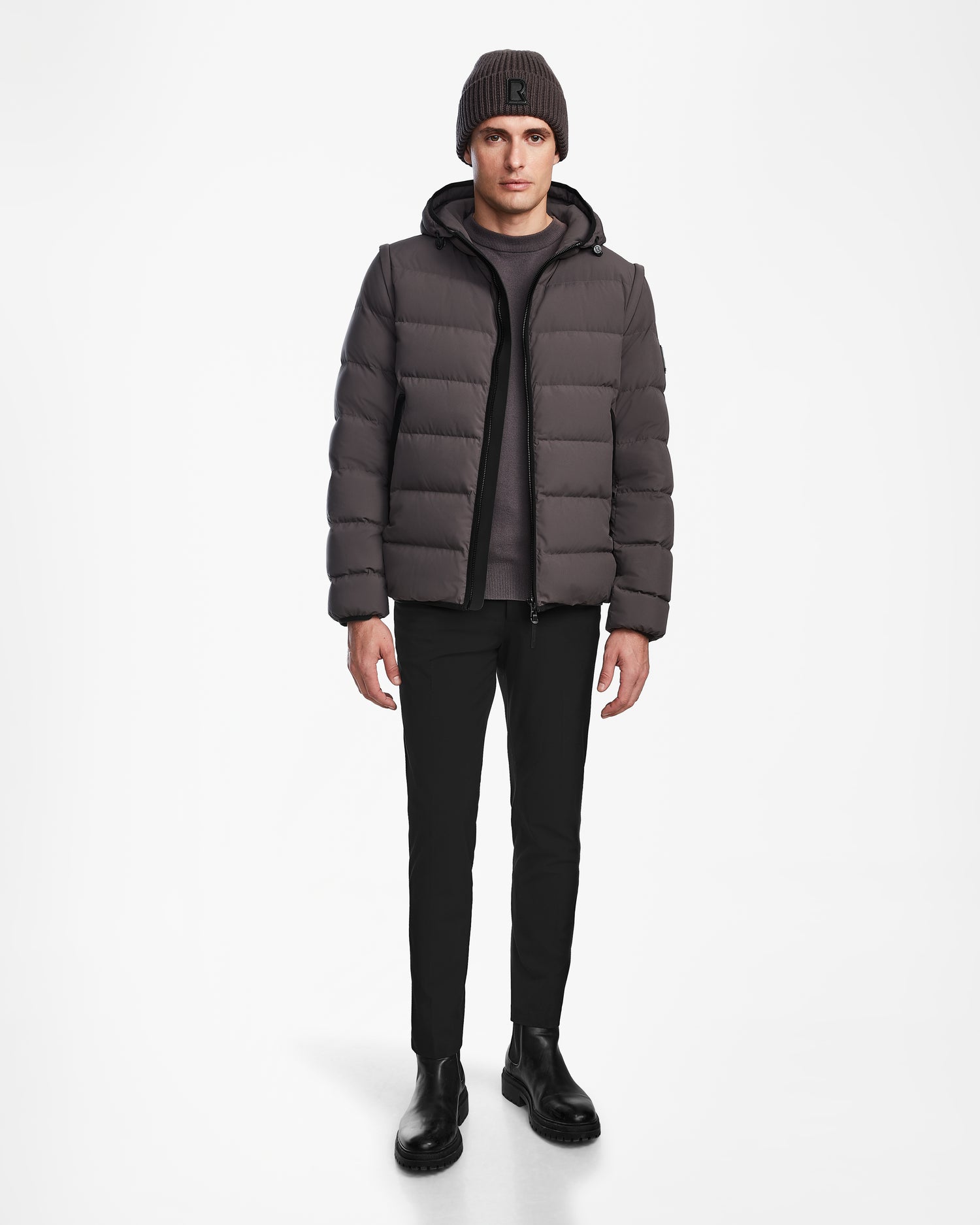 A man stands facing forward in the Rudsak DEMIAN M-R - M123504 Grey mens down puffer jacket, black pants, black boots, and a dark knit beanie, set against a plain white background.