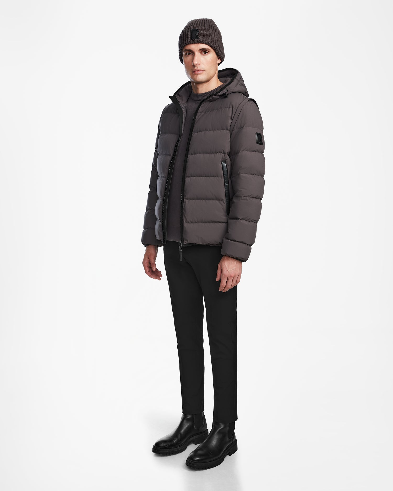 A person stands against a white background wearing the Rudsak DEMIAN M-R - M123504 Grey mens down puffer, styled with black pants, black boots, and a knitted gray beanie—crafted from sustainable recycled fabric.