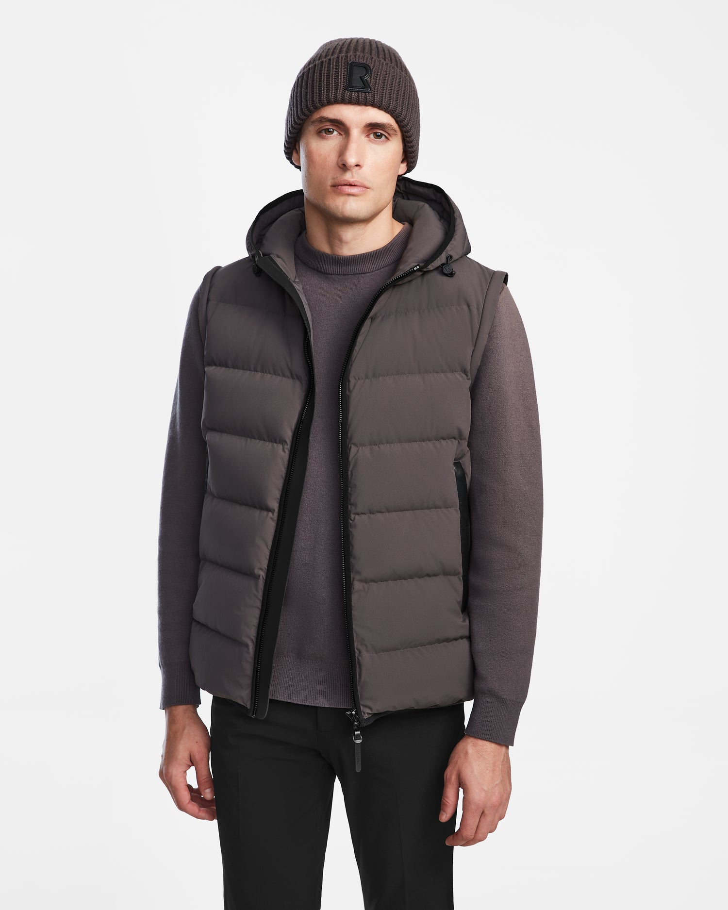 A man in the Rudsak DEMIAN M-R - M123504 Grey down puffer vest stands against a plain white background, paired with a matching gray sweater, black pants, and a gray knit beanie.