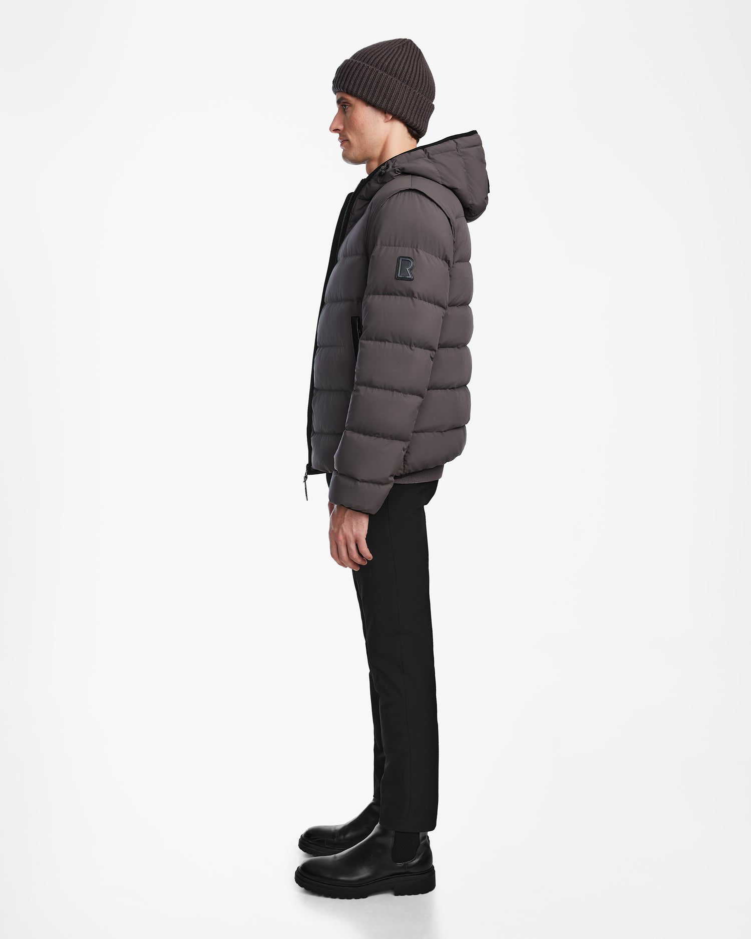 A man in profile wears the Rudsak DEMIAN M-R - M123504 Grey mens down puffer jacket with a hood, paired with black pants, black boots, and a dark knit beanie against a plain white background.