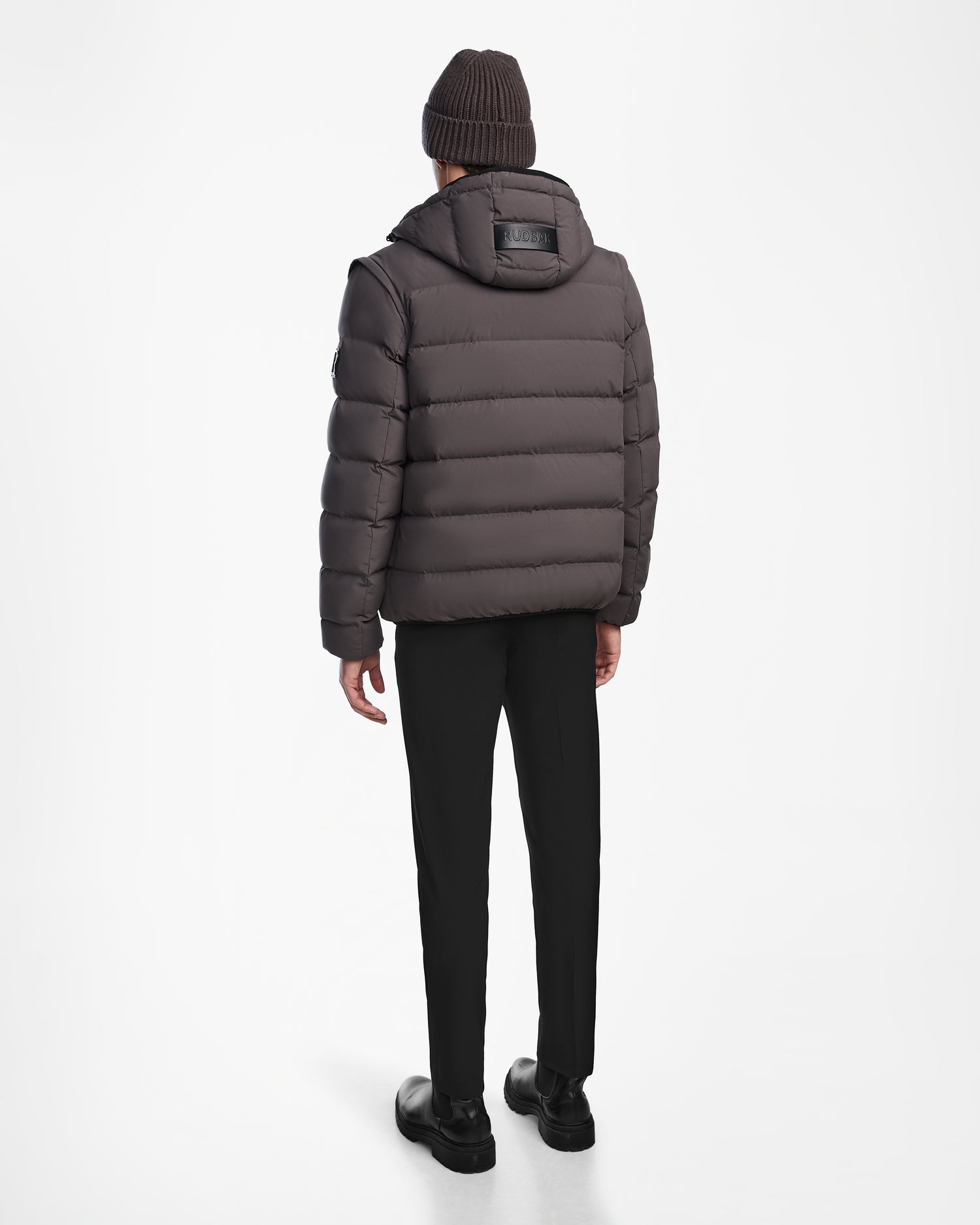 A person wearing a Rudsak DEMIAN M-R - M123504 Grey mens down puffer jacket stands facing away on a white background, styled with black pants, black shoes, and a brown knit beanie.