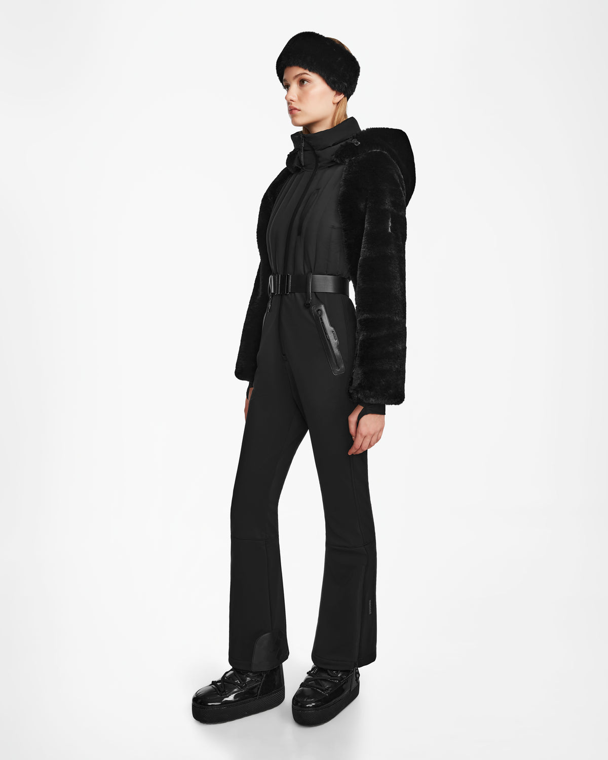 A woman models the Rudsak DESTINY - 8124727 Black ski suit, featuring a fur hooded jacket and matching pants with a wide belt, platform shoes, and a black fur hat, standing against a white background.