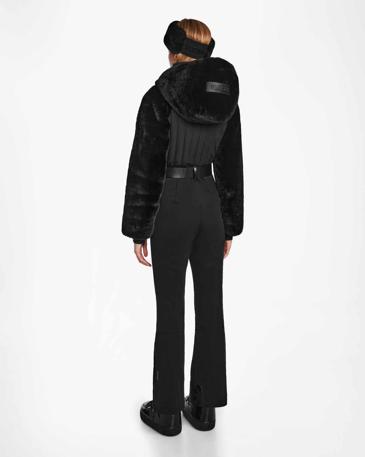 A woman wears the Rudsak DESTINY - 8124727 Black ski suit, styled with a fur hood, black boots, and earmuffs, standing facing away from the camera against a white backdrop. This womens suit offers warmth and style on the slopes.