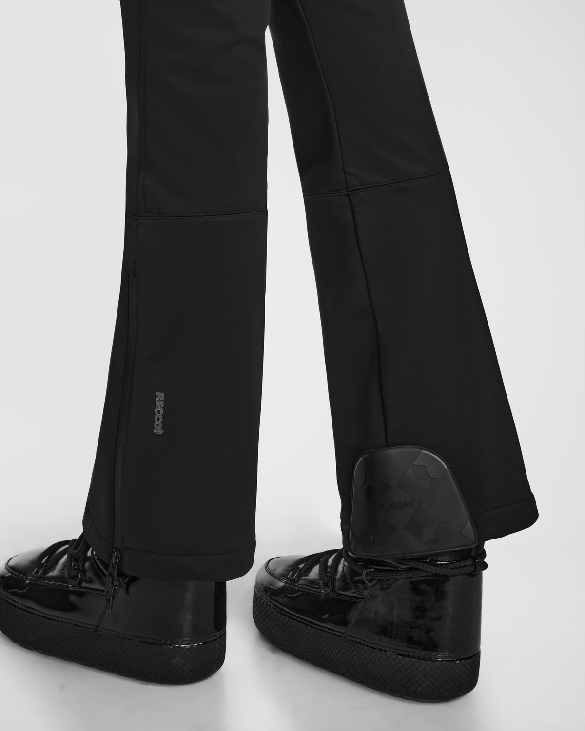 Close-up of a person wearing Rudsak DESTINY - 8124727 Black pants, with the pant leg slightly unzipped at the ankle to reveal a dark, angular device with RECCO rescue technology attached to shiny black sneakers.
