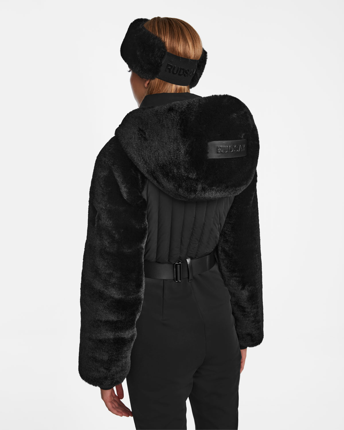A person with short hair is seen from behind wearing the Rudsak DESTINY - 8124727 Black faux fur hooded jacket, matching earmuffs, and belted pants. The jacket displays the Rudsak logo and discreet RECCO rescue tech for added mountain safety.