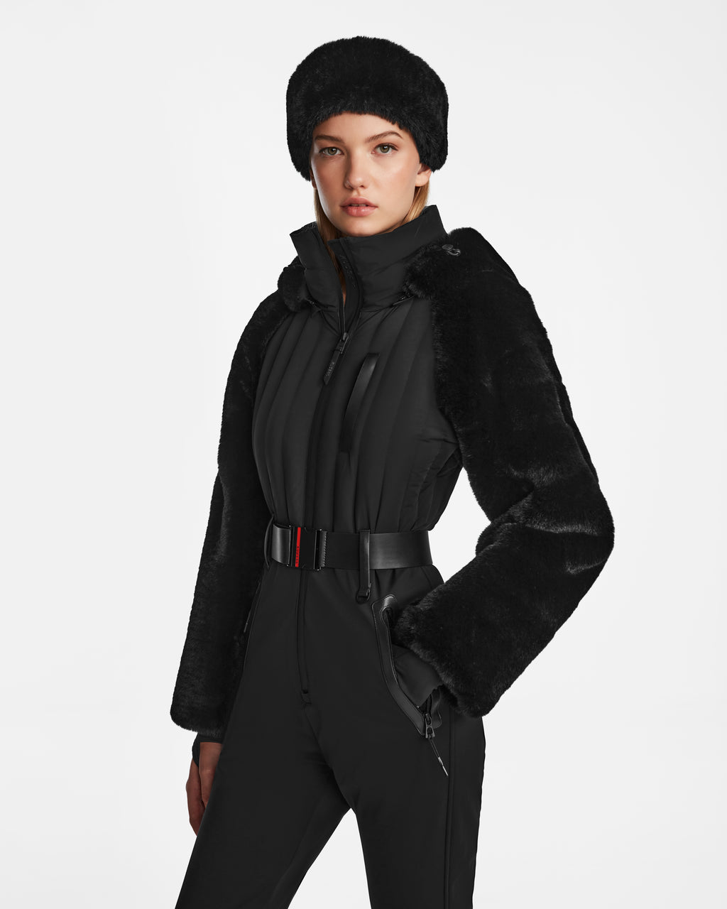 A woman models the Rudsak DESTINY - 8124727 Black: a black faux fur hat, a jacket with faux fur sleeves, and a belted women’s ski suit, all set against a plain white background.