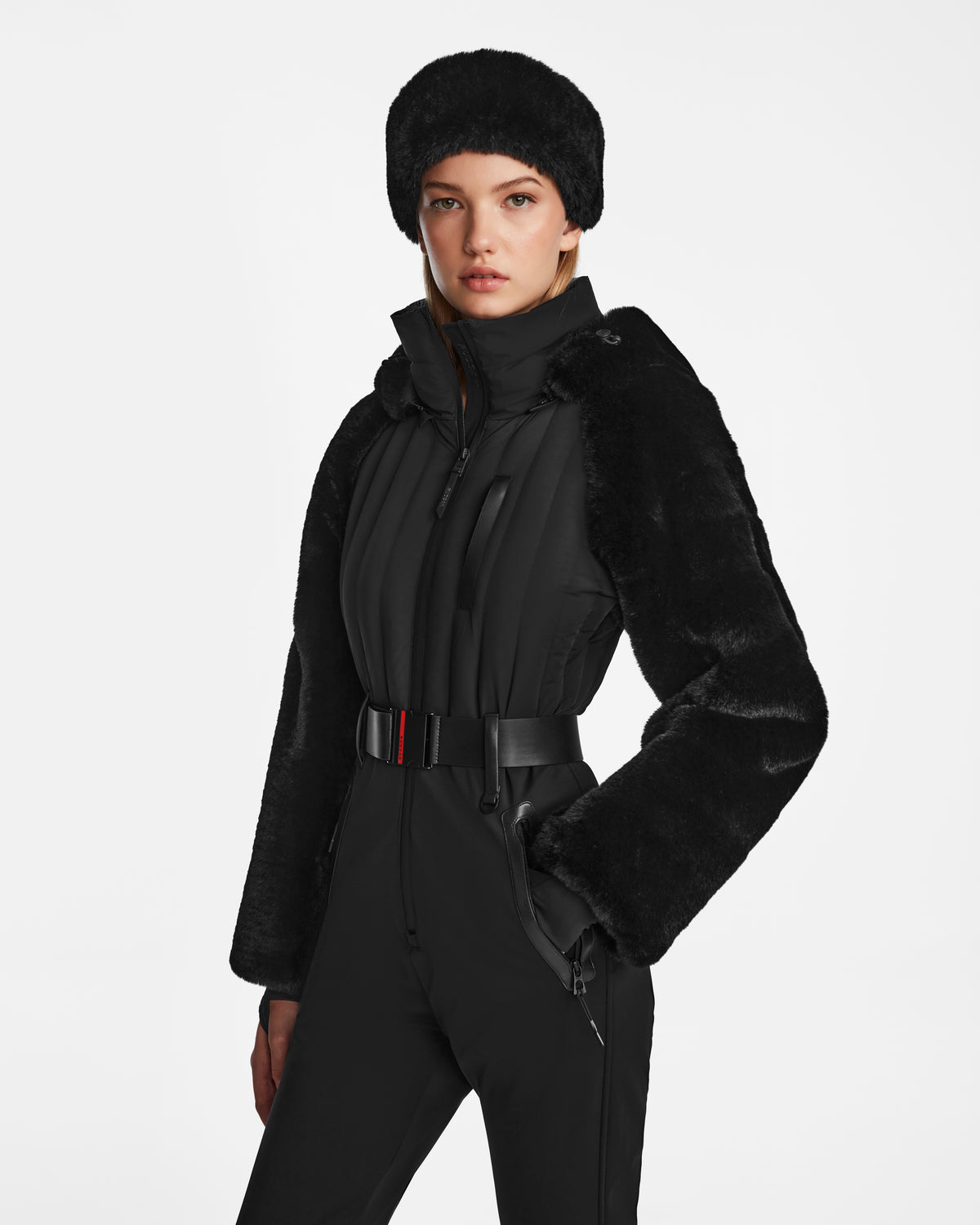A woman models the Rudsak DESTINY - 8124727 Black: a black faux fur hat, a jacket with faux fur sleeves, and a belted women’s ski suit, all set against a plain white background.