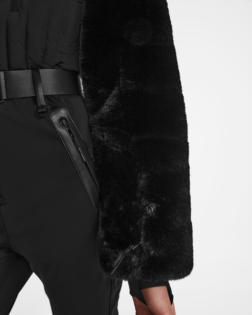 A model wears Rudsaks DESTINY - 8124727 Black womens ski suit, featuring black pants with zippered pockets, a wide black belt, and plush fur coat sleeve, shown waist down against a plain white background.