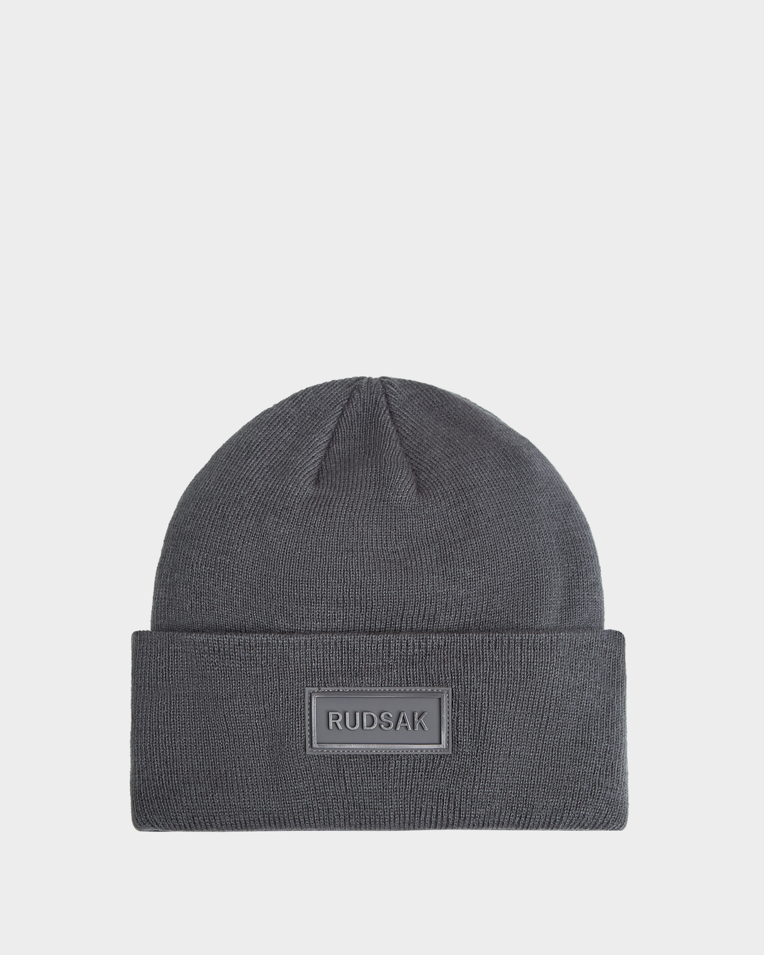 The Rudsak DILLION - U423501 Grey is a dark grey unisex knit hat with a folded brim and a front rectangular RUDSAK patch, shown on a plain white background—ideal for upgrading your winter style.