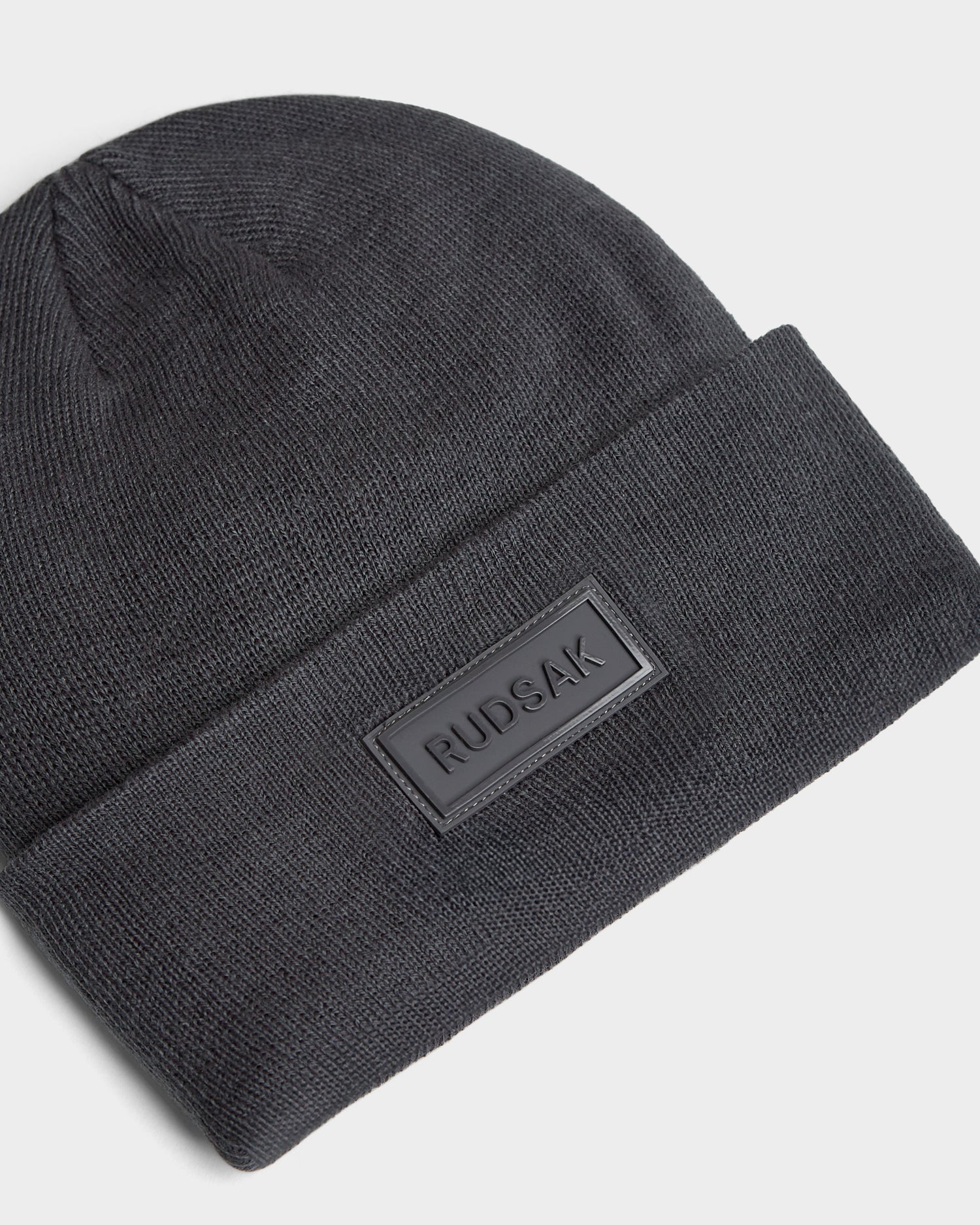 A grey unisex knit beanie named DILLION - U423501 by Rudsak features a folded brim and a rectangular black Rudsak patch on the front, laid flat on a white background—ideal for any winter wardrobe.