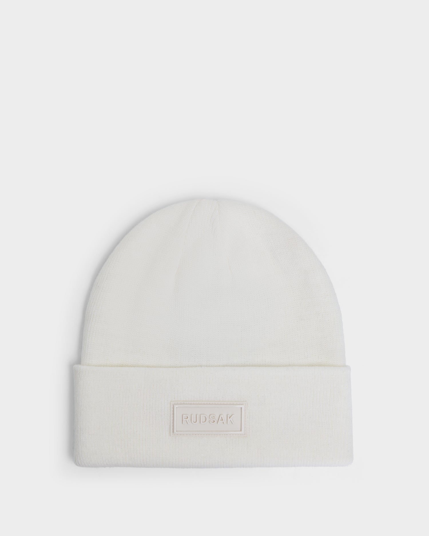 The DILLION - U423501 Optic White by Rudsak is a white unisex knit hat with a folded brim and a rectangular front patch embossed with "RUDSAK"—a classic winter accessory for any wardrobe.