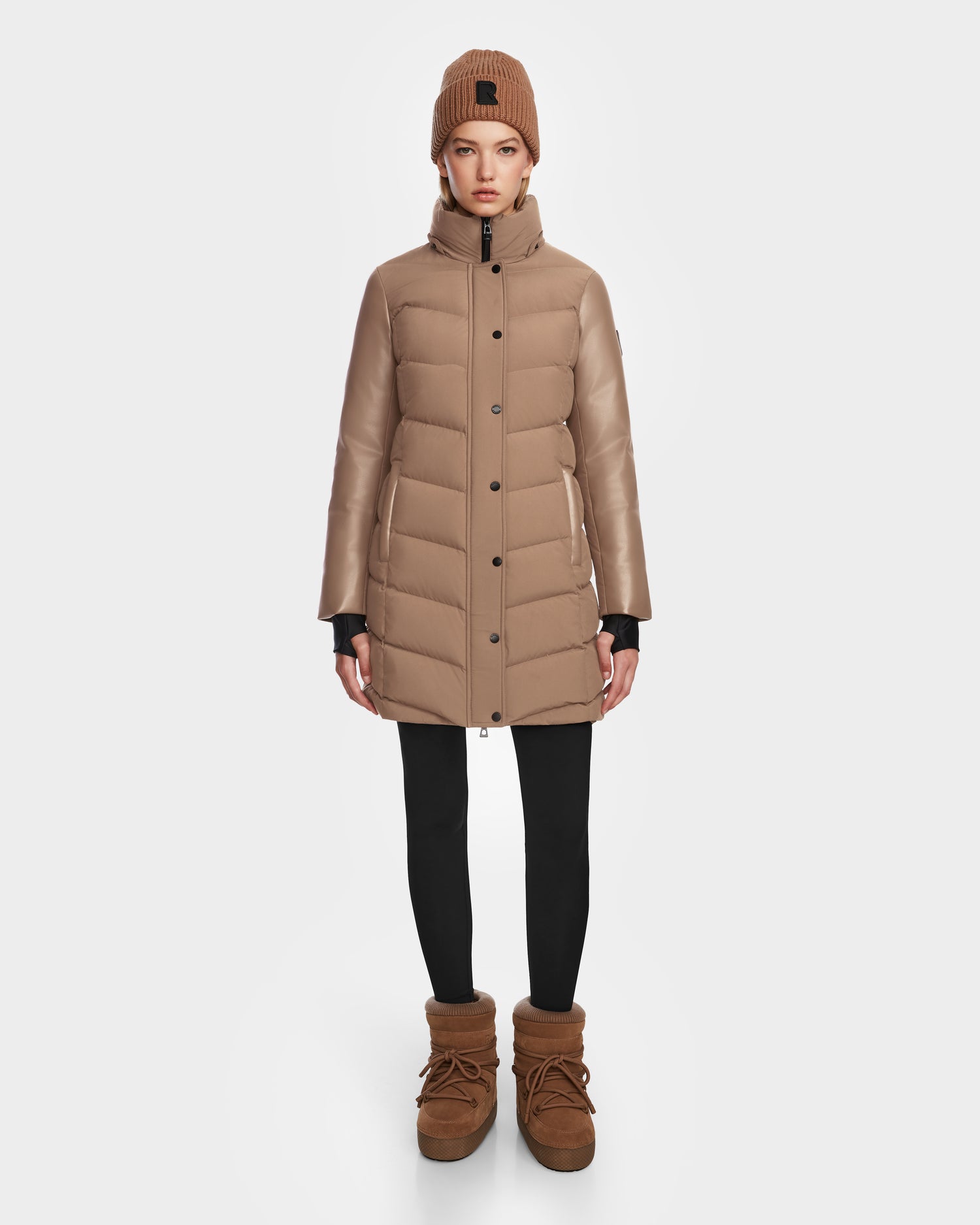 A woman faces forward wearing the Rudsak ELSA - L123535 Sand quilted down puffer coat, a matching beanie, black leggings, and brown winter boots against a plain white background.