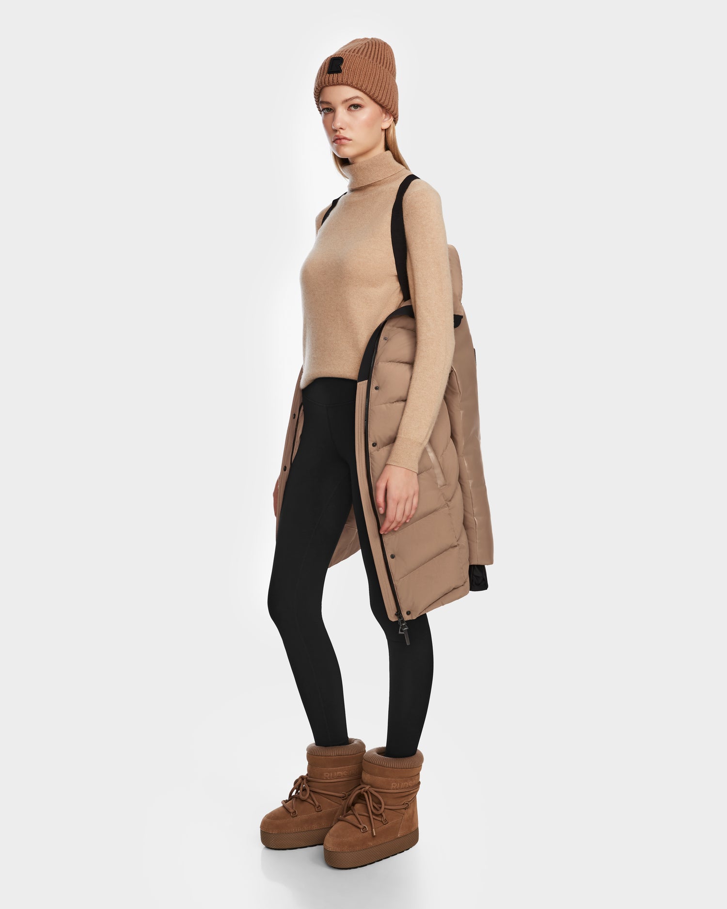 A woman stands against a plain white background, wearing the Rudsak ELSA - L123535 Sand sustainable winter jacket draped over one shoulder, paired with a tan turtleneck, black leggings, tan beanie, and tan boots.