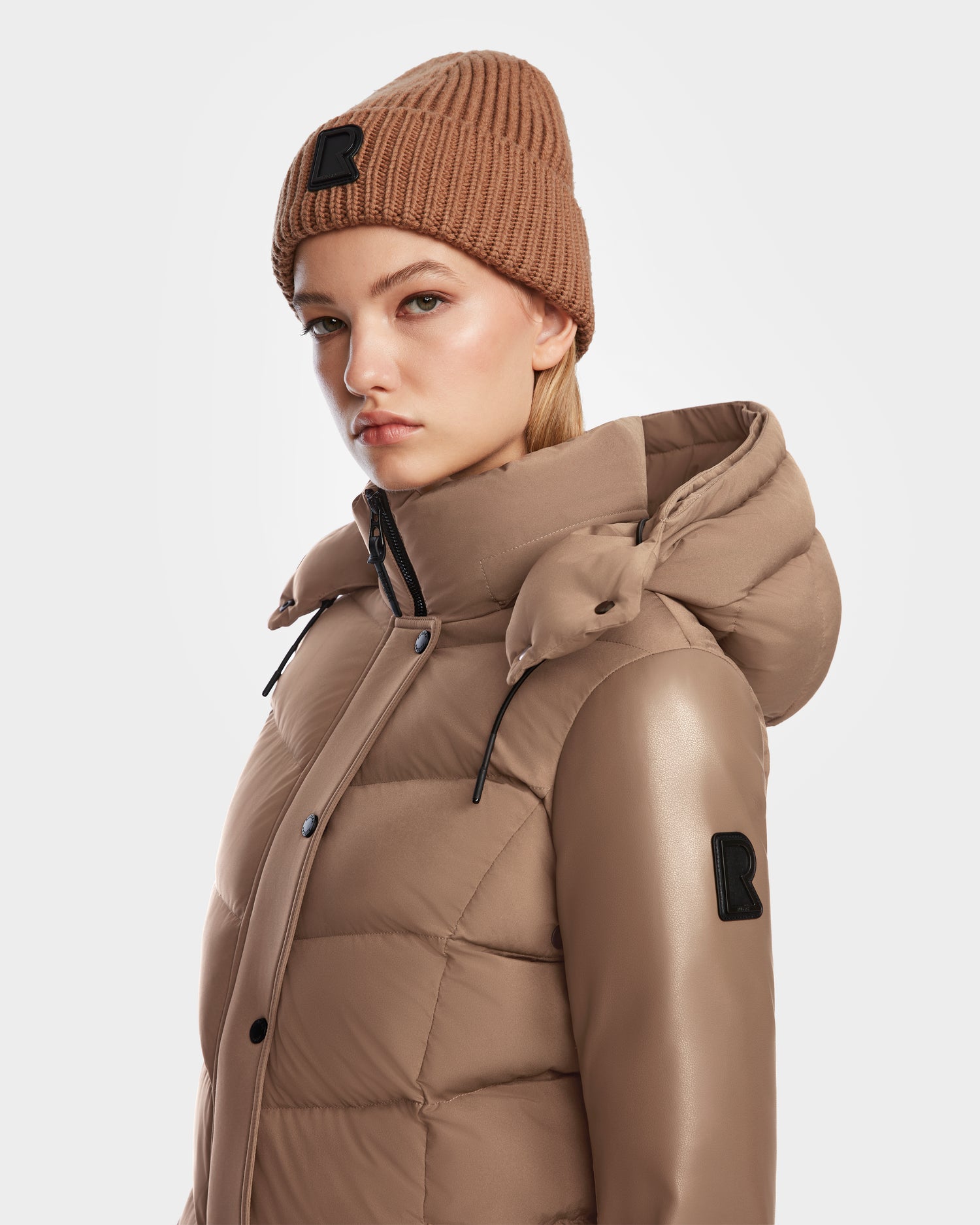 A woman wears the Rudsak ELSA - L123535 Sand quilted down puffer jacket with hood and a brown knit beanie, both featuring a black R logo patch, standing against a plain white background.