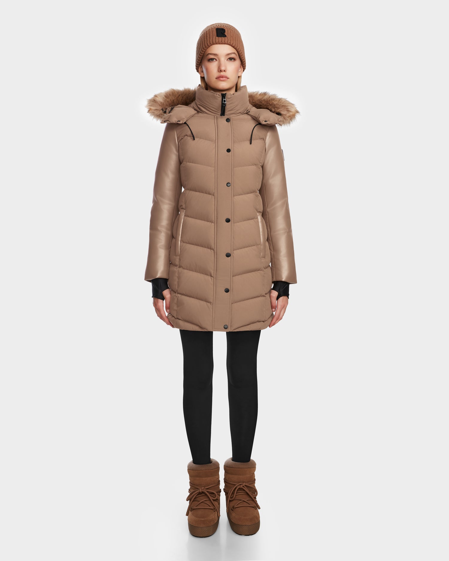A person stands facing forward in the Rudsak ELSA - L123535 Sand women’s quilted down puffer with a fur-lined hood, paired with black leggings, tan winter boots, and a matching tan knit beanie against a plain white background.