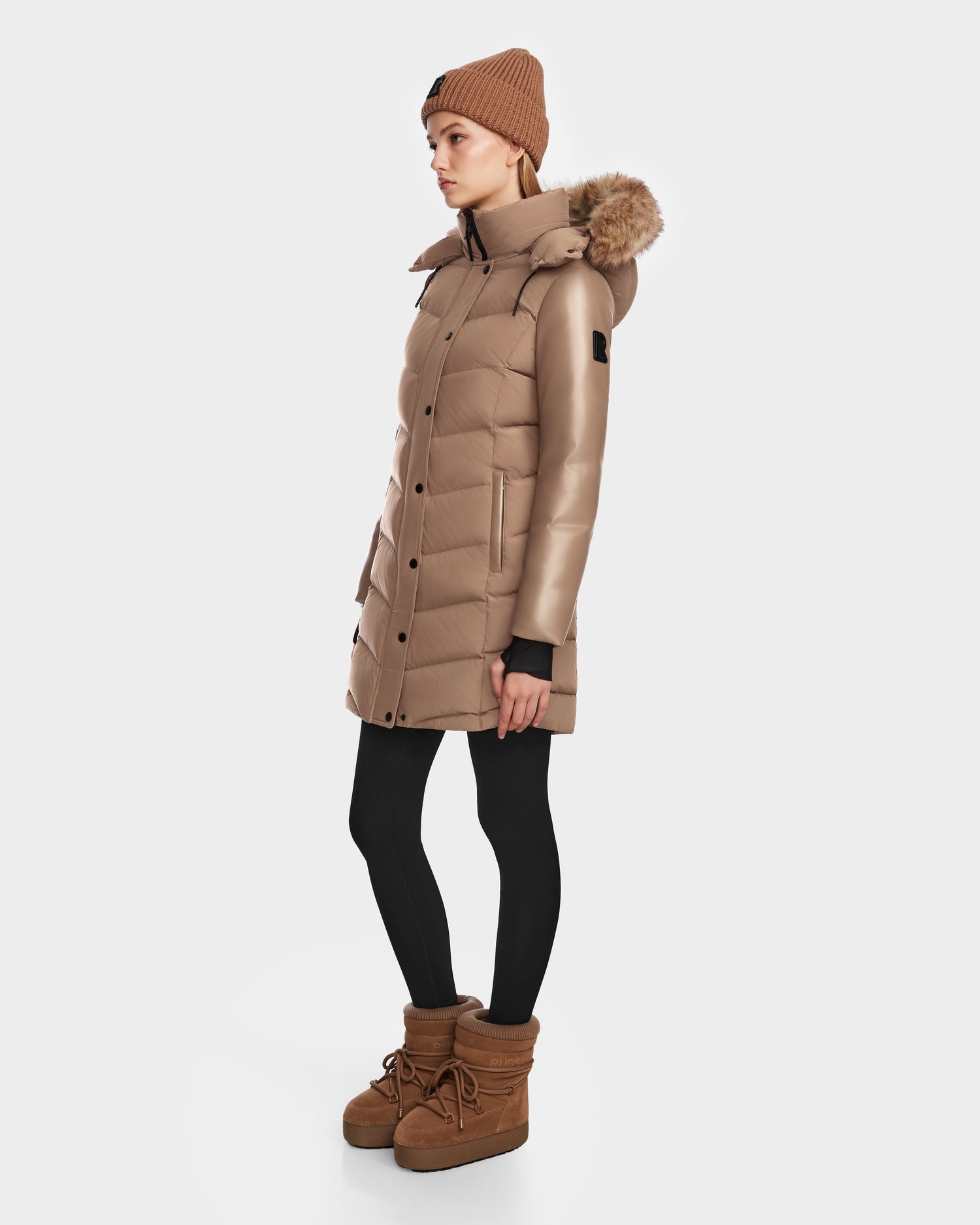 A person in profile wears the Rudsak ELSA - L123535 Sand women’s down puffer coat with a fur-lined hood, black leggings, tan winter boots, and a tan knit beanie against a plain white background.