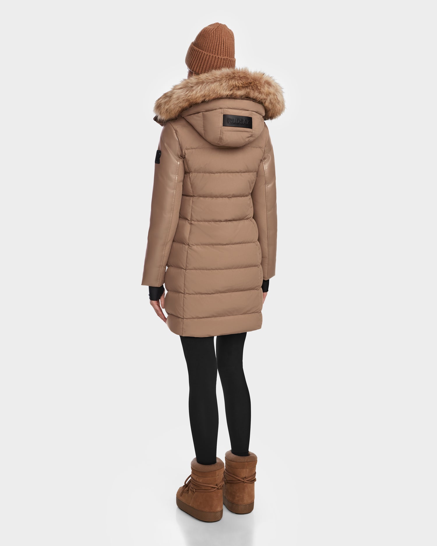 A person wearing the Rudsak ELSA - L123535 Sand women’s quilted down puffer with a fur-lined hood, black leggings, a brown beanie, and brown boots stands facing away against a plain white background.