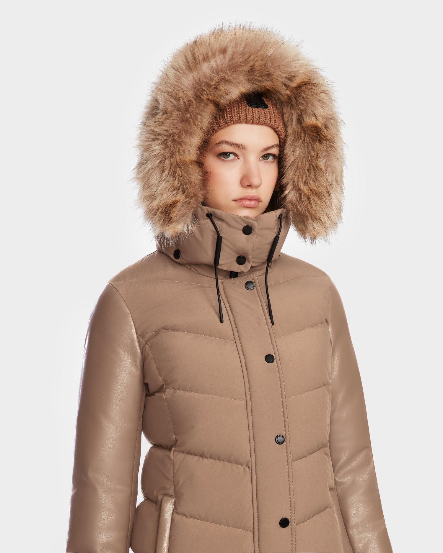 A person wears the Rudsak ELSA - L123535 Sand women’s quilted down puffer with a faux fur-lined hood and knit beanie, facing forward with a neutral expression against a plain white background.