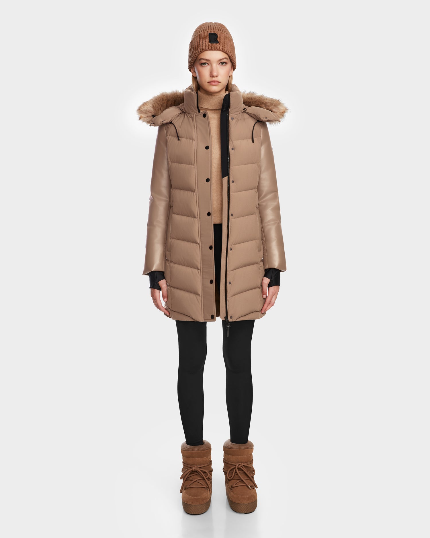 A woman stands facing forward against a plain white background, wearing the Rudsak ELSA - L123535 Sand puffer coat with a fur-trimmed hood, black leggings, tan winter boots, and a matching tan knit beanie.