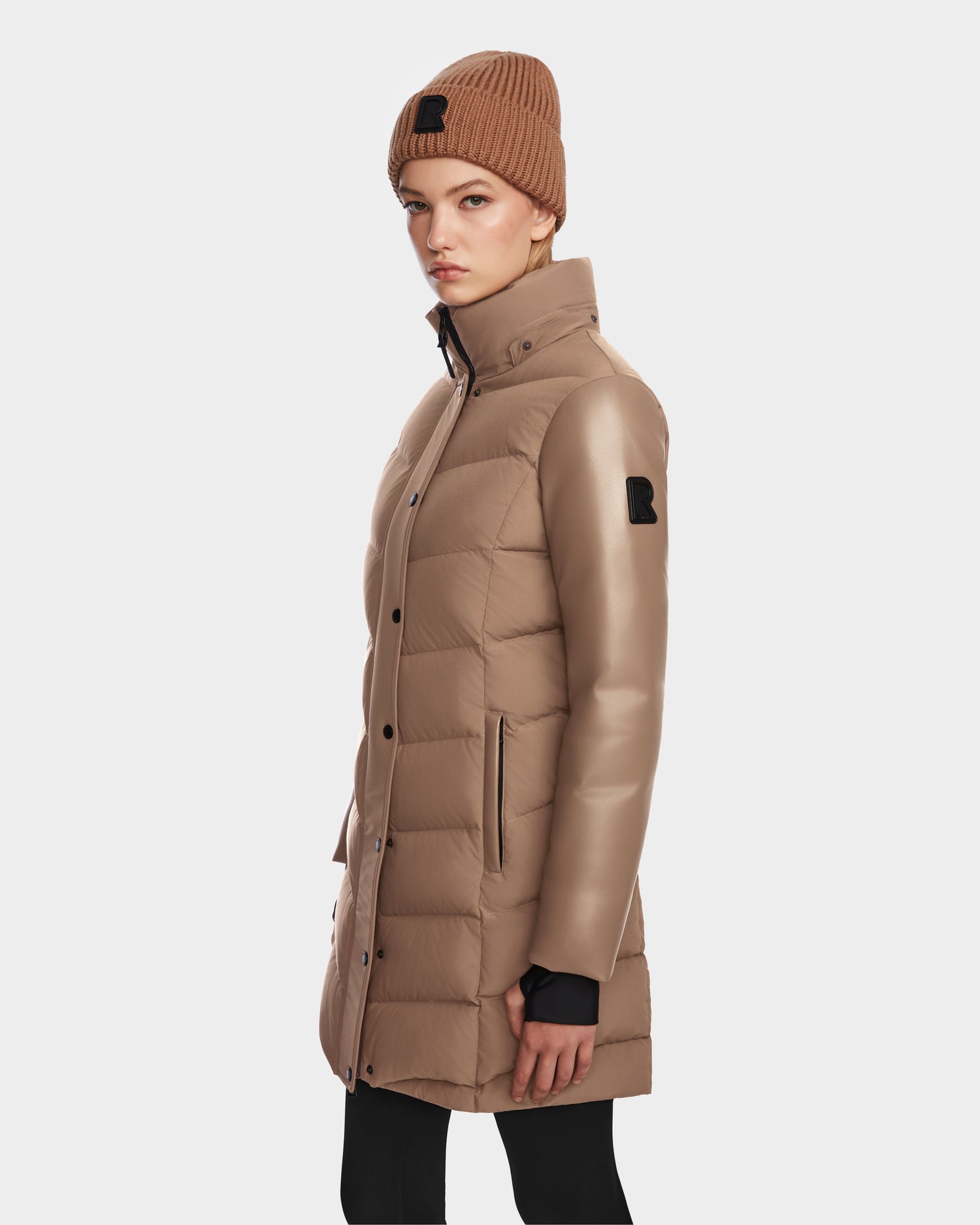 A woman in the Rudsak ELSA - L123535 Sand quilted down puffer with leather-look sleeves and a matching knit beanie stands sideways against a white background with a neutral expression.