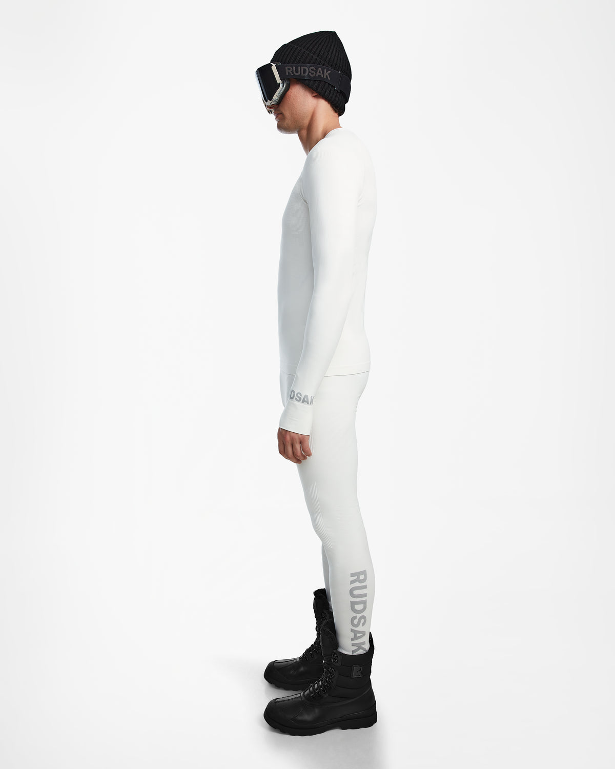 A person stands in profile wearing the Rudsak FRANC - 6524541 Iceland white fitted men’s thermal set, black winter boots, beanie, and ski goggles against a plain white background—ideal as a ski base layer.