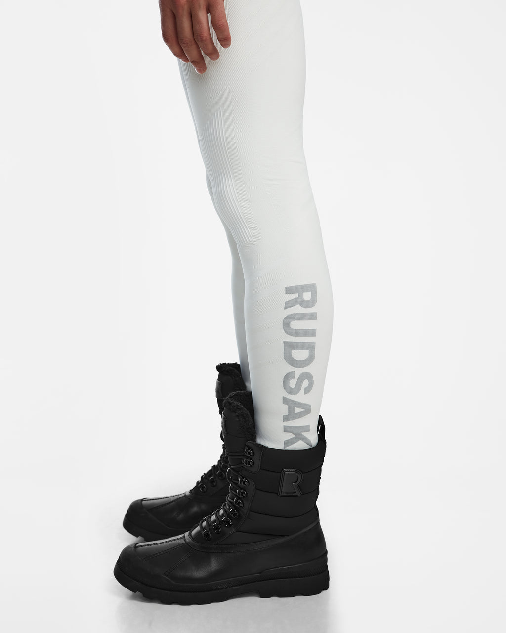A person wearing the Rudsak FRANC - 6524541 Iceland white stretch underlayer leggings with RUDSAK printed vertically on the side and black lace-up winter boots, standing against a white background.