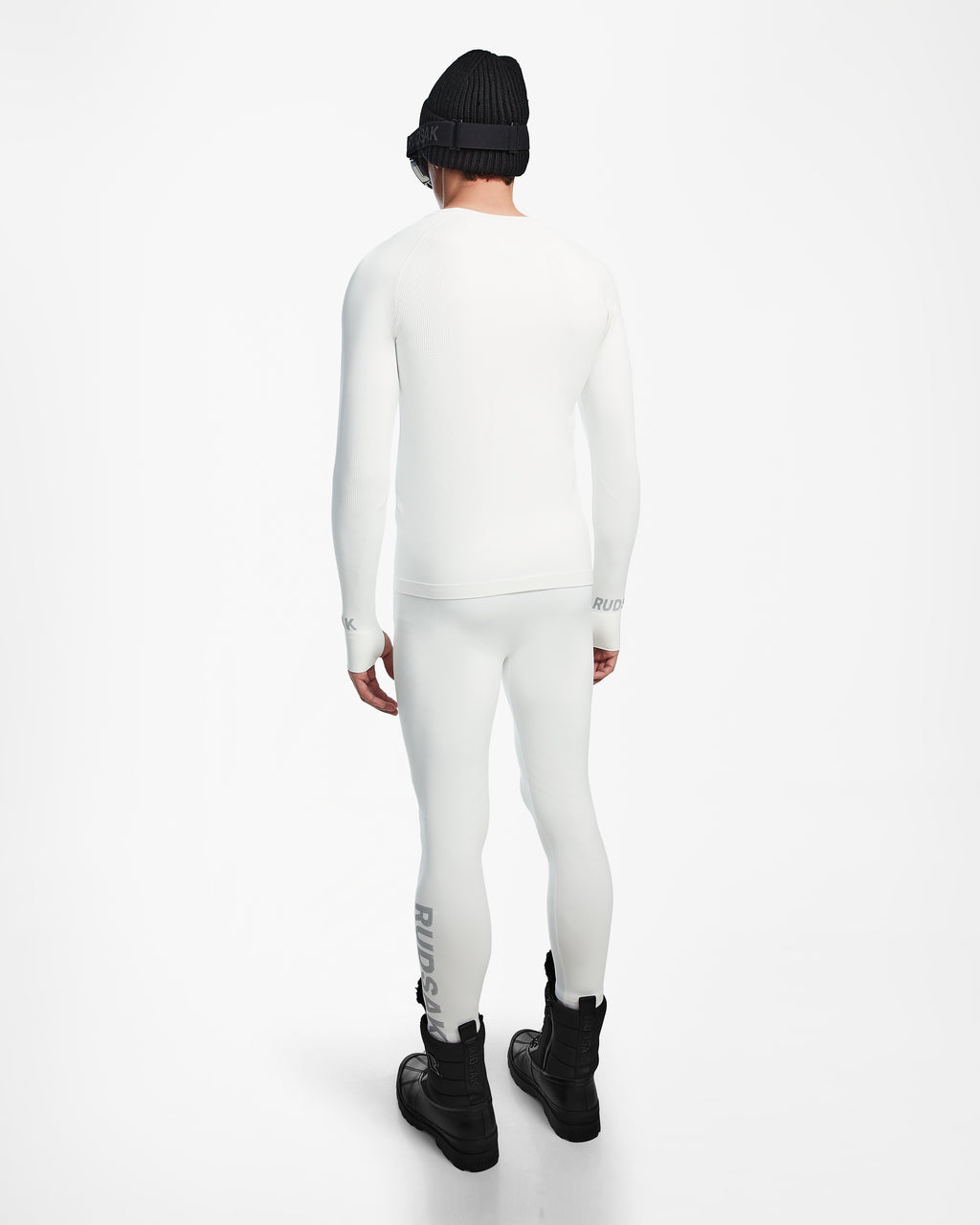 A person faces away, wearing the Rudsak FRANC - 6524541 Iceland fitted white long-sleeve ski base layer and leggings, paired with black boots, a black beanie, and sunglasses against a plain white background.