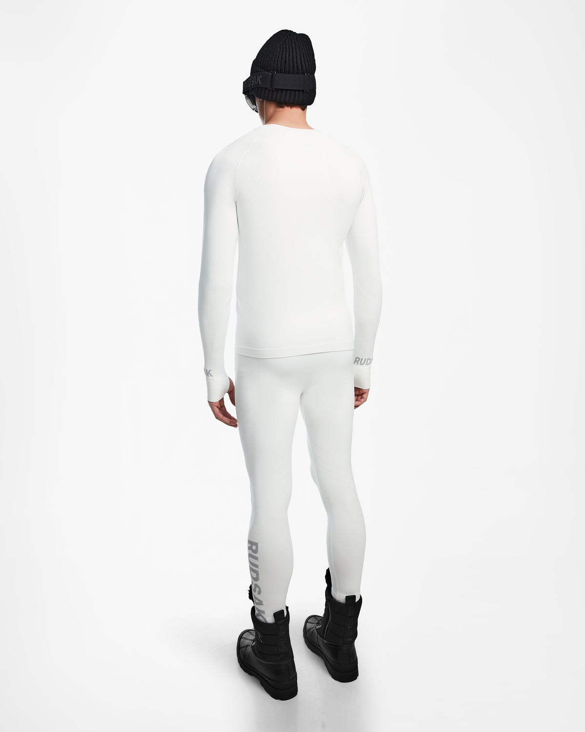 A person faces away, wearing the Rudsak FRANC - 6524541 Iceland fitted white long-sleeve ski base layer and leggings, paired with black boots, a black beanie, and sunglasses against a plain white background.