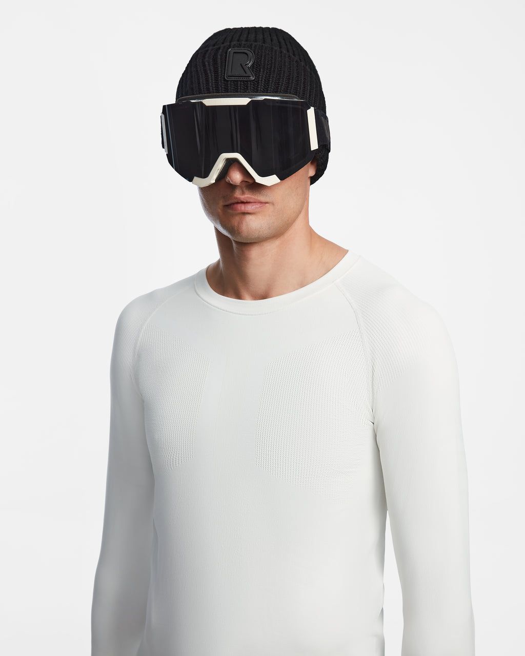 A man models the Rudsak FRANC - 6524541 Iceland black knit beanie, large black ski goggles with white accents, and a fitted white ski base layer against a plain white background.