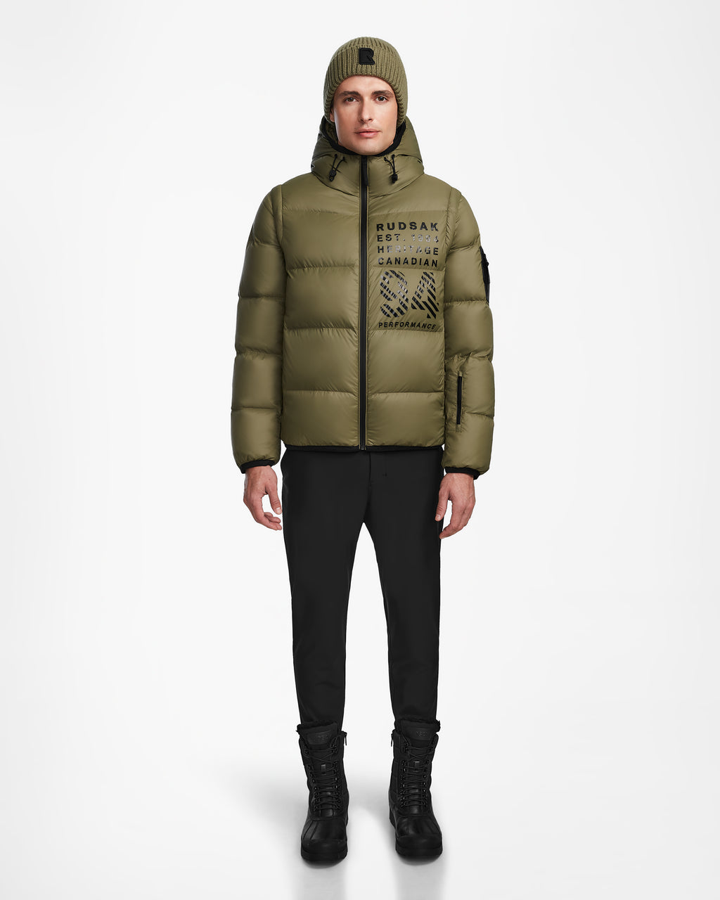 A person stands facing forward against a plain white background, wearing the Rudsak HANS - 6124518 Loden Green men’s down ski jacket with RECCO rescue technology, paired with a matching beanie, black pants, and black boots. The wind- and water-resistant jacket provides both style and protection in harsh conditions.
