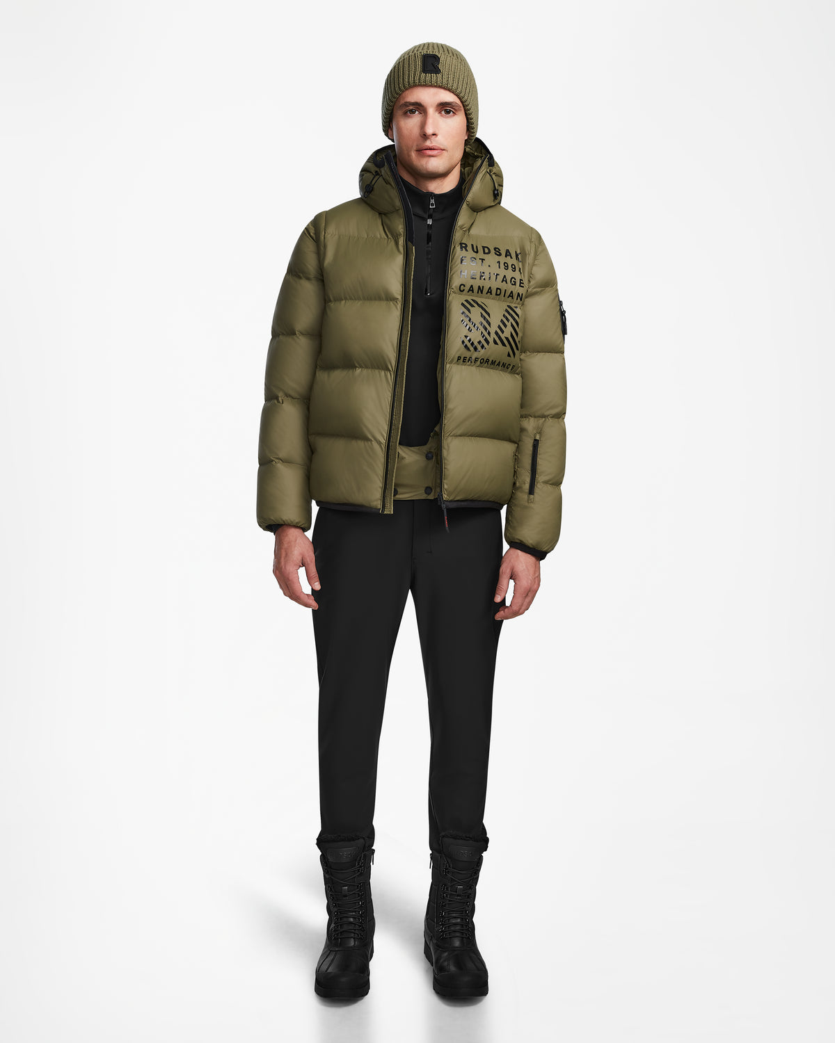 A man wearing the Rudsak HANS - 6124518 Loden Green puffer jacket with RECCO rescue technology, along with black pants, black boots, and a green beanie, stands facing forward against a plain white background.