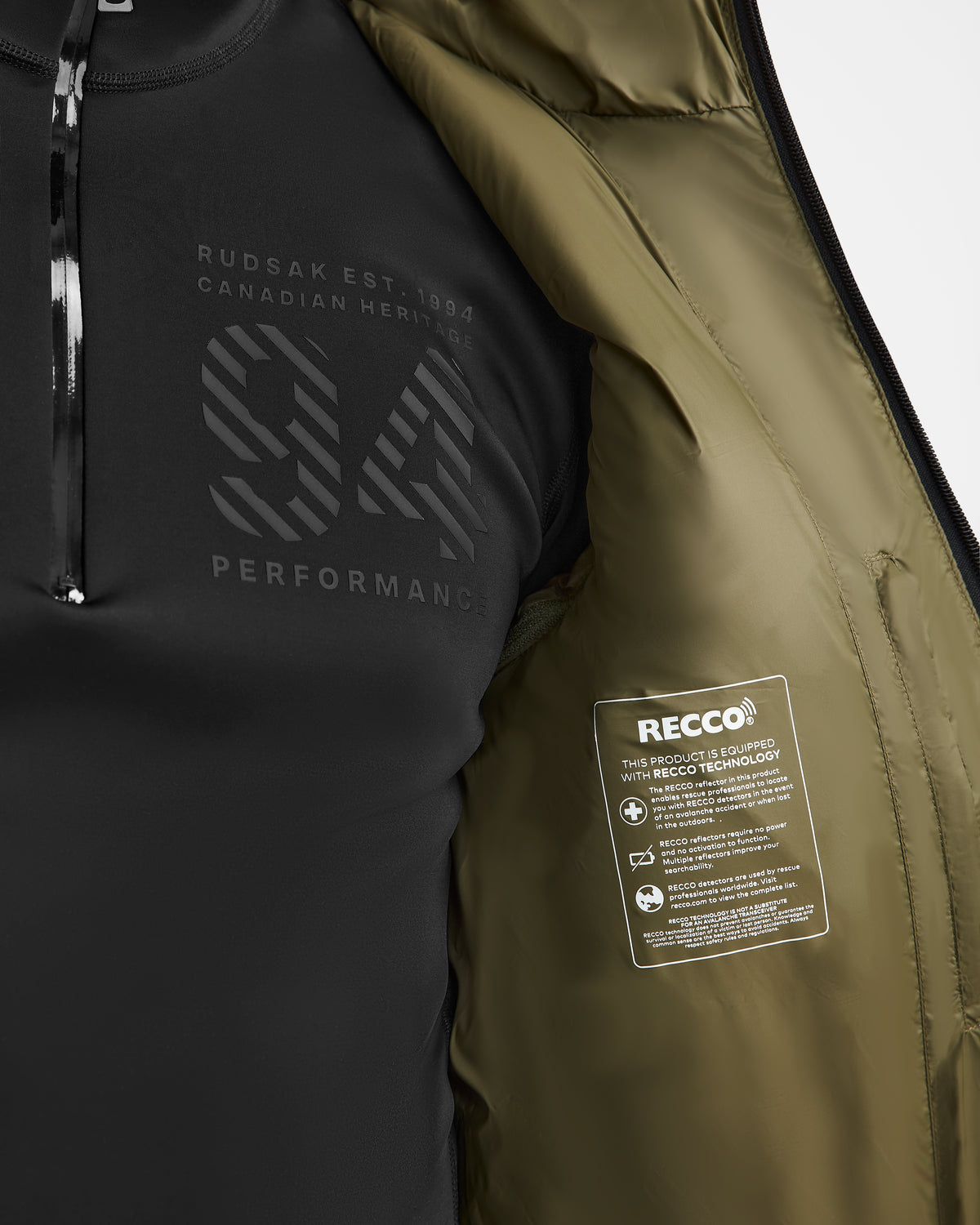 A close-up of a person wearing a black shirt with “94 PERFORMANCE” printed on it. The interior of the Rudsak HANS - 6124518 Loden Green men’s down ski jacket is visible, showcasing the RECCO rescue technology information label and highlighting its wind- and water-resistant features.