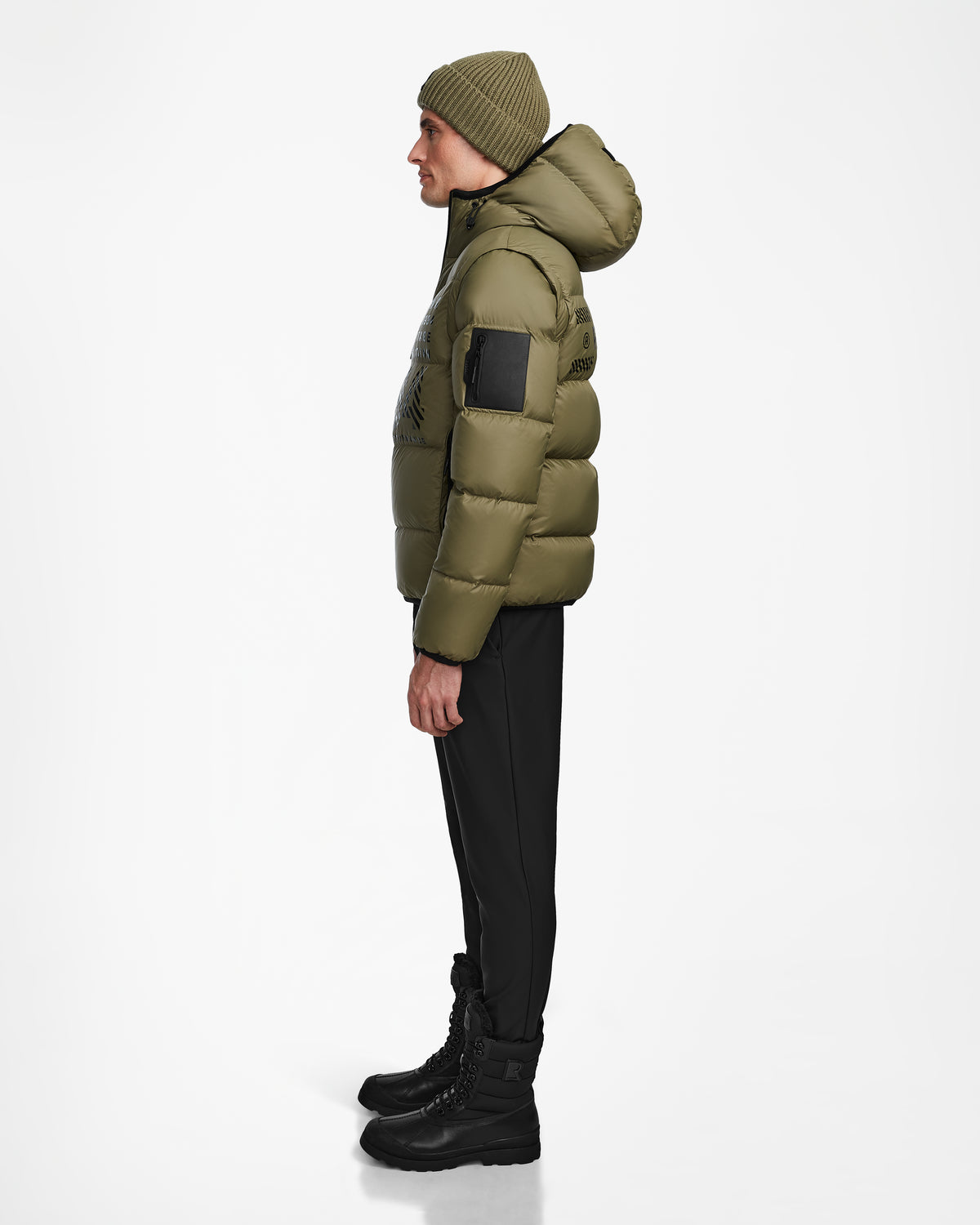 A person stands in profile against a plain white background, dressed in the Rudsak HANS - 6124518 Loden Green men’s down ski jacket—an olive green, wind and water-resistant style—paired with black pants, black boots, and a matching knit beanie.