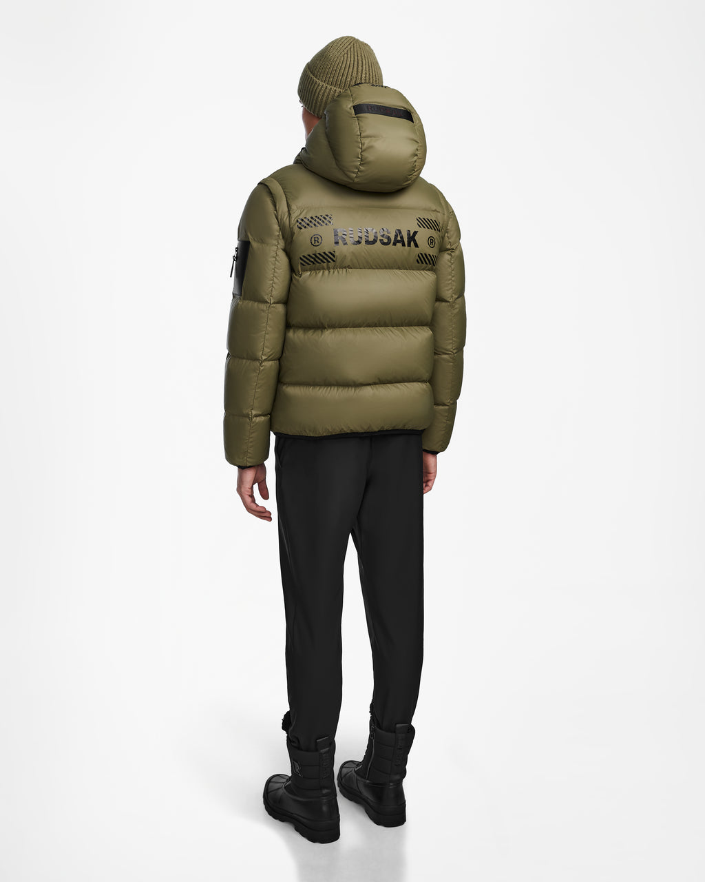 A person is standing with their back to the camera against a plain white background, wearing the Rudsak HANS - 6124518 Loden Green men’s down ski jacket—an olive green wind and water-resistant piece featuring RECCO rescue technology—paired with black pants, black boots, and a matching green beanie.