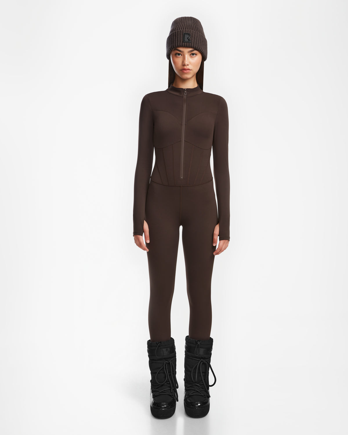 A person stands facing forward in the Rudsak IVY - 8524599 Espresso fitted womens stretch bodysuit with a zip-up front, paired with a matching knit beanie and black snow boots against a plain white background.