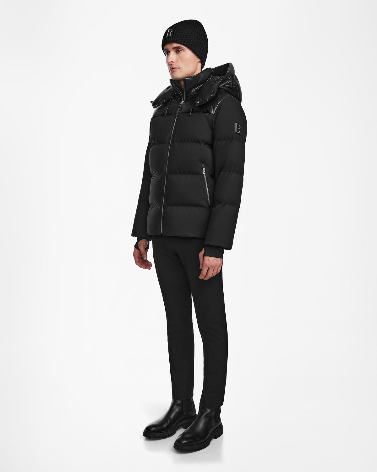 A person stands against a white background wearing Rudsak’s JACK - M124507 Black men’s down puffer jacket with a hood, along with a black knit beanie, fitted black pants, and black boots.