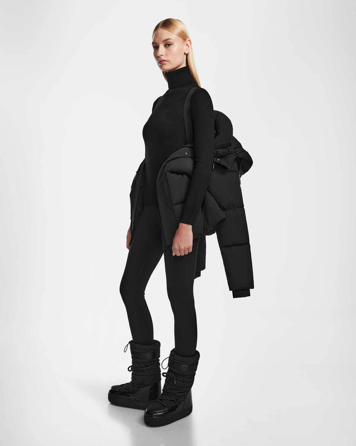 A woman stands against a white background wearing Rudsaks JOON - 8124728 Black turtleneck, black leggings, black winter boots, and holding an RDS-certified down puffer jacket over one shoulder.