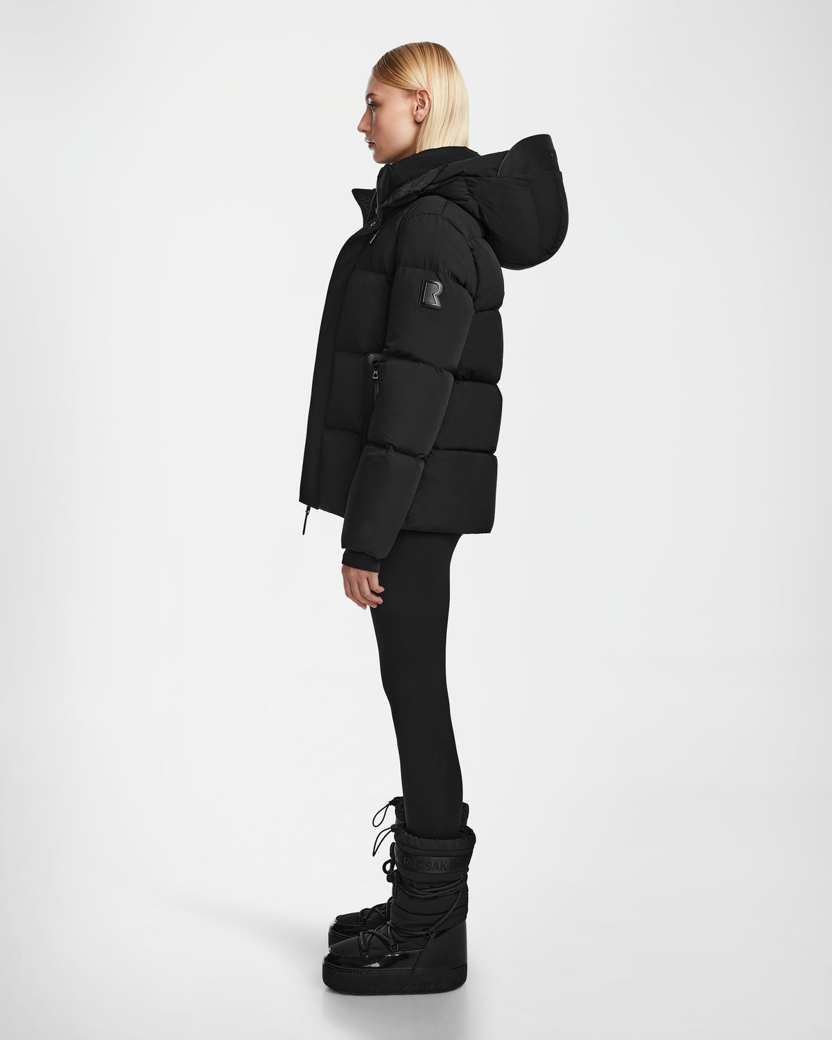 A woman with straight blonde hair, wearing the Rudsak JOON - 8124728 Black down puffer jacket, black leggings, and winter boots, stands in profile against a plain white background.
