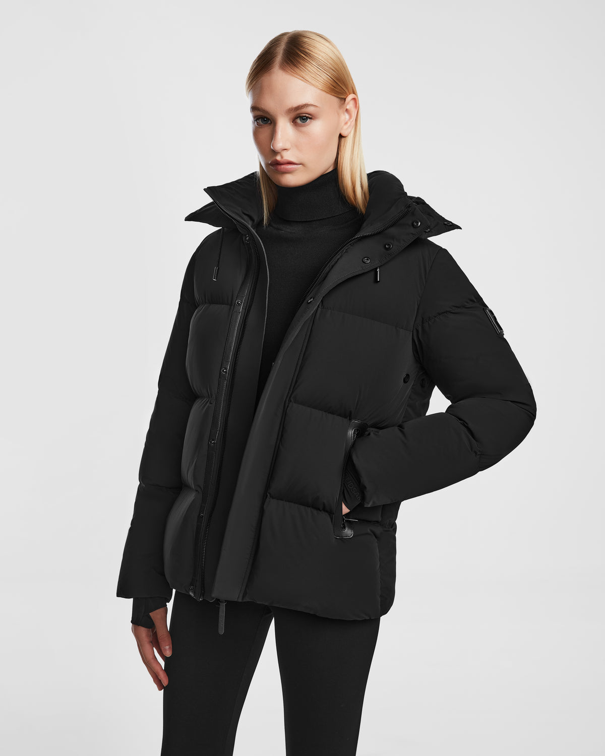 A woman with straight blonde hair wears the Rudsak JOON - 8124728 Black RDS-certified down puffer jacket over a black turtleneck and pants, standing against a plain white background.