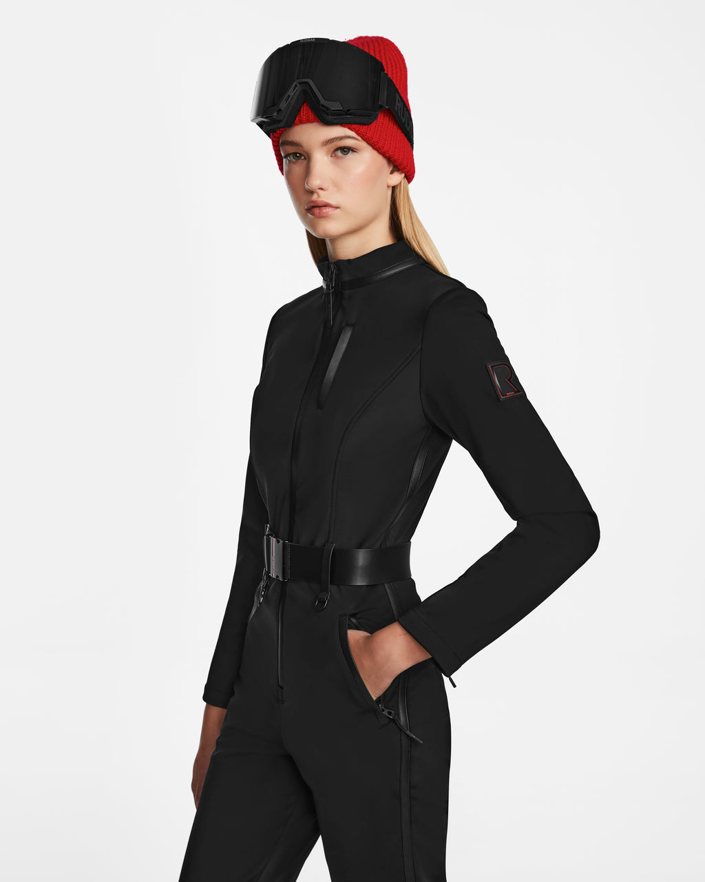A woman wears the Rudsak JOSIE - 8124817 Black one-piece ski suit with a belted waist and RECCO rescue tech, paired with a red beanie and black ski goggles, standing against a plain white background.