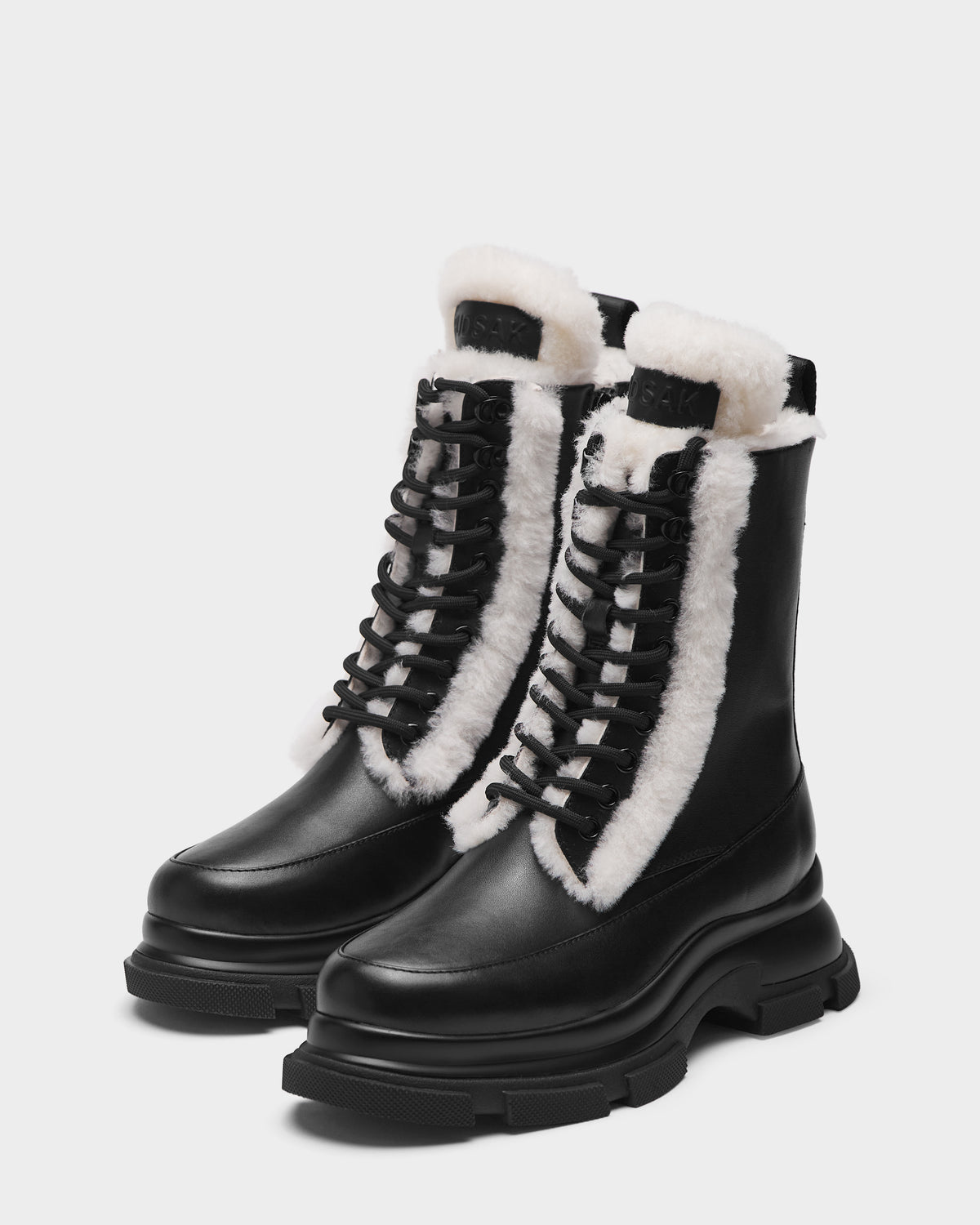 The Rudsak LANA S - 8224537 Black boots feature chunky soles, black mid-calf lace-up design, and visible white shearling lining at the tongue and sides, set against a plain light background.