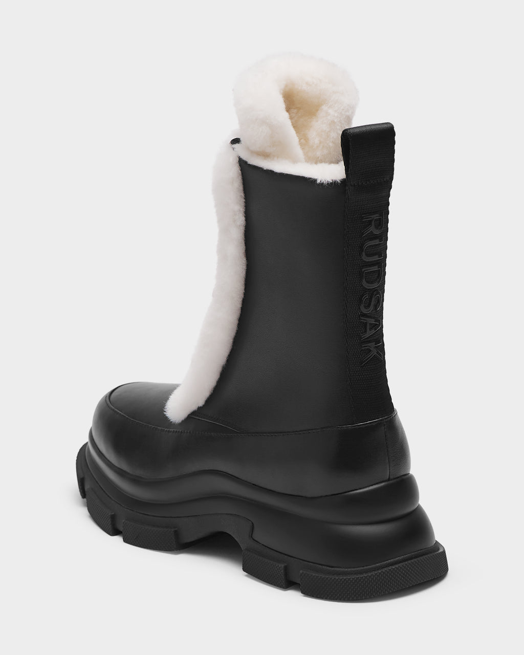 The Rudsak LANA S - 8224537 Black is a women’s black leather shearling boot with a chunky sole, visible white shearling lining, and a rear pull tab embossed with RUDAK—perfect for winter, shown on a plain light background.