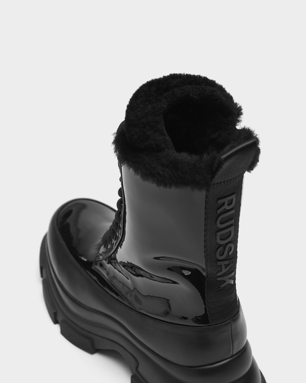 A close-up of the Rudsak LANA - 8223506 women’s winter boot in black, featuring a glossy finish, thick treaded sole, shearling lining, and Rudsak shown vertically on a fabric tab at the back.
