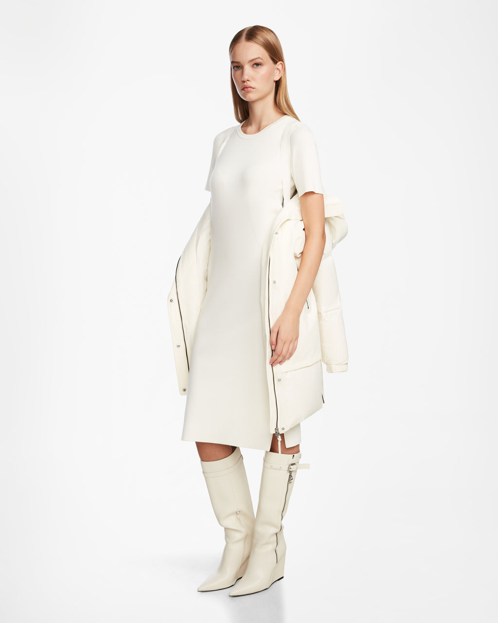 A woman stands against a plain white background, wearing the Rudsak LAYAN - 8124576 Iceland vegan leather winter coat draped over her shoulders, paired with a simple knee-length white dress and knee-high white boots.