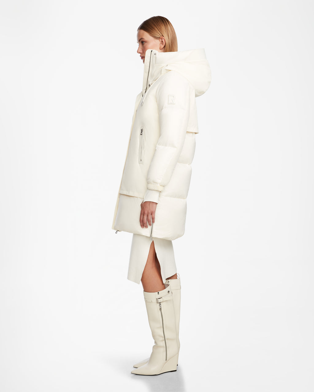 A woman stands in profile against a white background wearing the Rudsak LAYAN - 8124576 Iceland off-white down parka with hood, paired with matching knee-high vegan leather boots and a white front-slit skirt.