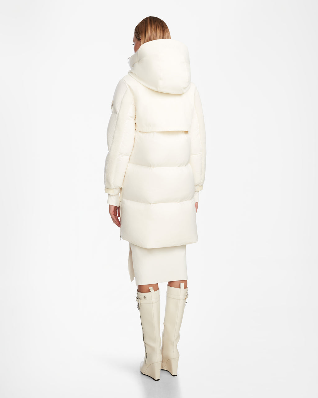 A person faces away, wearing the Rudsak LAYAN - 8124576 Iceland womens down parka with a large hood, paired with a white skirt and tall white zippered boots, set against a plain white background.