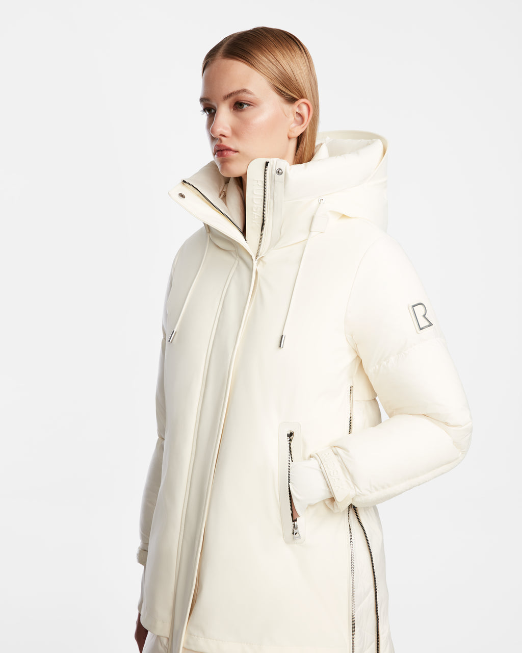 A woman with straight blonde hair models the Rudsak LAYAN - 8124576 Iceland off-white womens down parka, featuring zippered pockets, a hood, and a logo patch on the upper sleeve, standing against a plain white background.