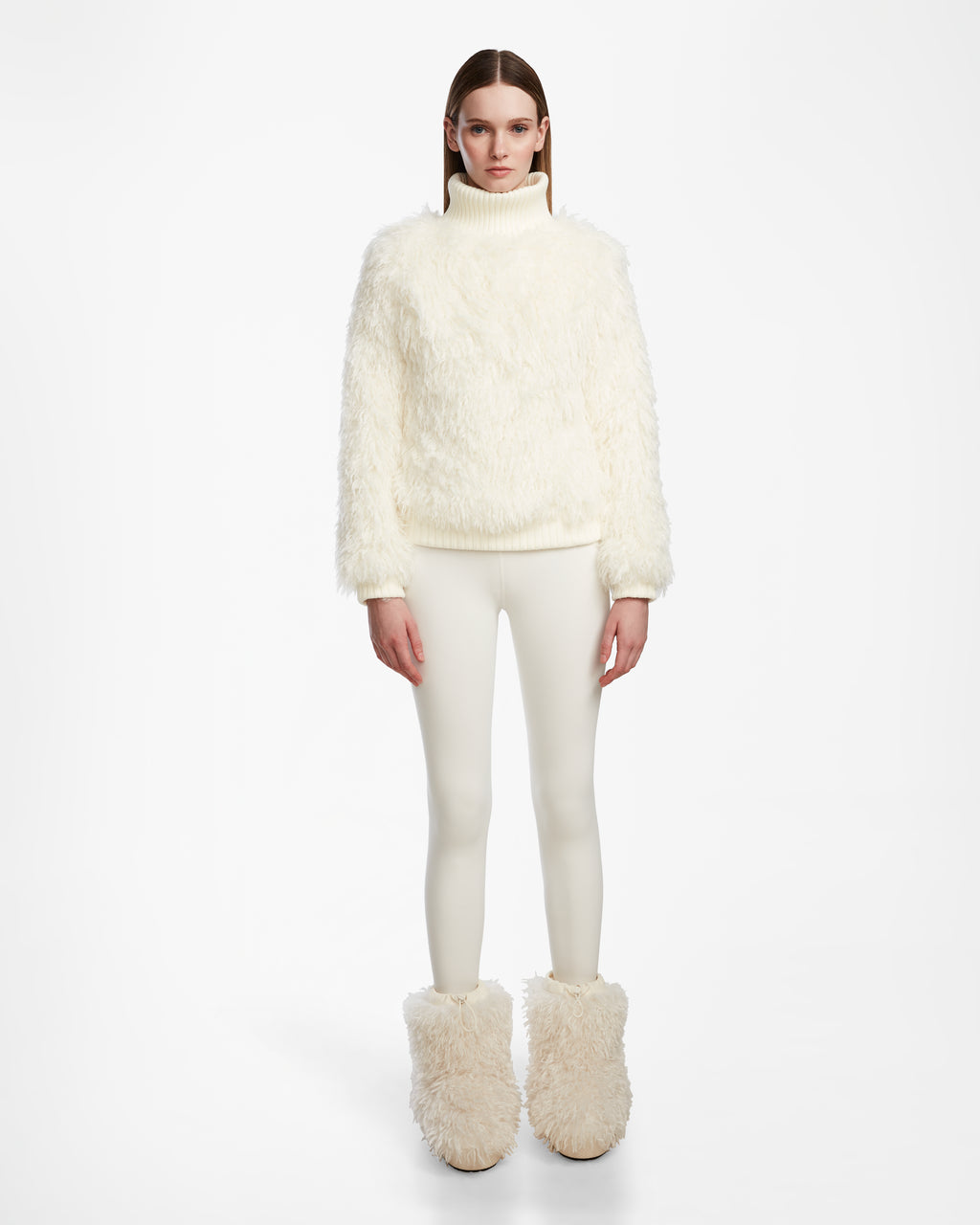 A woman faces forward in the Rudsak LAYLA M - 8524600 Iceland faux Mongolian fur turtleneck, paired with fitted white pants and furry boots, capturing après-ski style against a plain white background.
