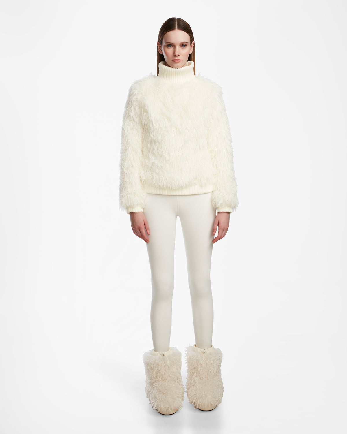 A woman faces forward in the Rudsak LAYLA M - 8524600 Iceland faux Mongolian fur turtleneck, paired with fitted white pants and furry boots, capturing après-ski style against a plain white background.